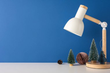 Minimalist bakış açısı İskandinav masa sahnesinde modern bir lamba ve dekoratif mini Noel ağaçları mavi bir duvara yaslanmış, sıcacık kış titreşimlerini yakalıyor.