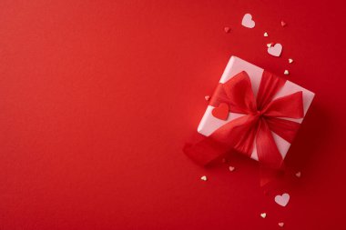 Çok güzel paketlenmiş kırmızı bir hediye kutusu. Etrafı kalp şeklinde dekorasyonlarla çevrili bir kurdele. Kırmızı arka planda sevgi ve kutlama var.