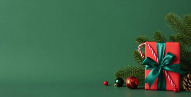 Yeşil kurdeleli ve şeker kamışlı kırmızı bir hediye Noel süsleri ve çam dallarının yanında duruyor. Minimalist tasarım rahat ve şenlikli bir tatil atmosferini çağrıştırıyor.