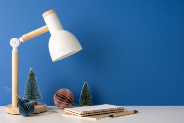 Mavi bir duvara karşı ahşap bir lamba, defter ve şenlikli Noel süslemelerinin yer aldığı bir yan manzara. Kış temalı ya da İskandinav tarzı tasarımlar için ideal.