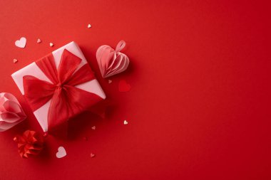 Güzel paketlenmiş kırmızı kurdeleli bir Sevgililer Günü hediyesi kırmızı arka planda kırmızı kağıt kalpler ve konfetilerle çevrilidir. Romantik kutlamalar için ideal.