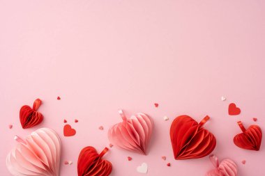 Kırmızı ve pembe kağıt kalpler açık pembe arka planda dağılır ve Sevgililer Günü için romantik ve şenlikli bir ortam yaratır.