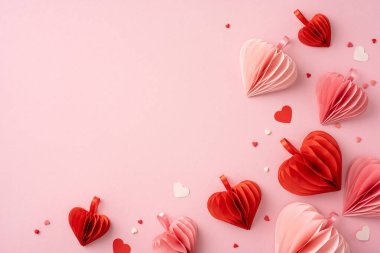 Hassas kırmızı ve pembe kağıt kalpler yumuşak pembe bir arka plana dağılmış, Sevgililer Günü için romantik ve şenlikli bir atmosfer yaratıyor.