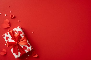 Kalp desenli kağıda sarılmış kırmızı kurdeleli, küçük kırmızı kalplerle süslenmiş bir hediye. Sevgililer Günü kutlamaları, aşk ve romantizm için ideal.