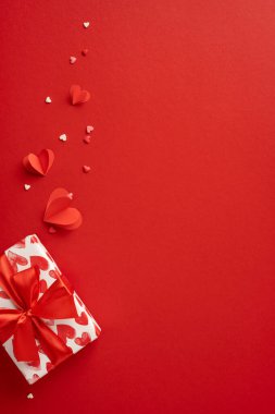 Dikey romantik bir Sevgililer Günü sahnesi. Güzel paketlenmiş bir hediye kutusu, kırmızı kalpler ve kırmızı arka planda kağıt süslemeler. Sevgi ve sevgiyi ifade etmek için mükemmel.