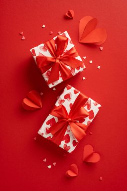 İki dikey güzel paketlenmiş Sevgililer Günü hediyesi kırmızı kalp desenleri ve parlak kırmızı bir zemin üzerinde kurdeleler kağıt kalplerle çevrili.