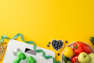 Meyveler, sebzeler, dambıllar, bir tartı ve mezura içeren parlak ve renkli kompozisyon, sağlıklı bir yaşam tarzını ve dengeli beslenmeyi simgeliyor.