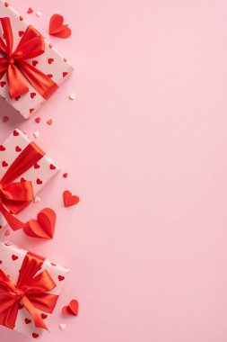 Sevgililer Günü dikey hediyeleri kalp desenli kağıda sarılmış kırmızı fiyonklu yumuşak pembe arka planda, küçük kalp şekilleriyle süslenmiş.