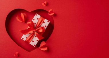 Çok güzel paketlenmiş bir hediye kutusu içinde kalpler ve kırmızı bir kurdele, canlı kırmızı bir zemin üzerine kurulmuş, Sevgililer Günü kutlamaları ve romantik kutlamalar için mükemmel.