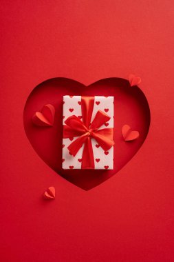 Kırmızı kurdeleli dikey bir hediye kutusu kırmızı bir kalp şeklinde duruyor. Aşkı ve sevgililer gününü simgeliyor.