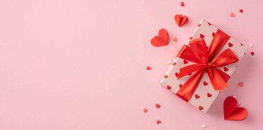 Güzel paketlenmiş bir Sevgililer Günü hediyesi pembe arka planda uzanan canlı kırmızı kurdeleli, dağınık kalp şekilli dekorasyonlarla çevrili romantik ve şenlikli bir atmosfer yaratıyor.