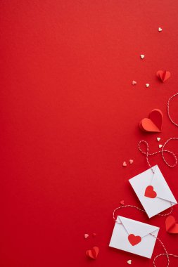 Dikey olarak kırmızı bir sevgililer günü arka planında kağıt kalpler, küçük zarflar ve kalp konfetileri romantik ve şenlikli bir atmosfer yaratıyor.