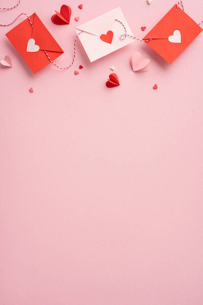 Красные и белые вертикальные конверты с вырезанными сердцами и разбросанными бумажными сердцами на мягкой розовой поверхности создают романтическое настроение ко Дню Святого Валентина