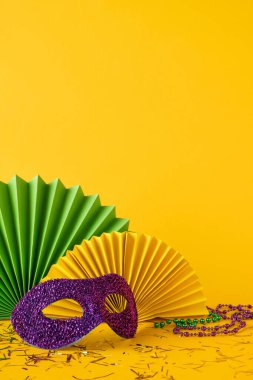 Renkli Mardi Gras yan görünüm dekorasyonları parlak bir maske, yelpaze ve parti boncukları ile gösterilen canlı sarı arka planda konfeti ve kutlama unsurları yer alır.