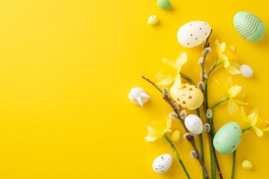 Renkli boyanmış Paskalya yumurtaları ve taze bahar çiçekleri yaratıcı bir şekilde sarı bir zemine dizilmiş.