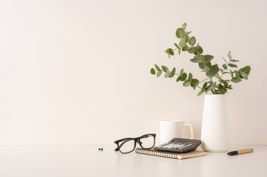 Gözlük, hesap makinesi, defter, kalem ve okaliptüs içeren minimalist çalışma alanı
