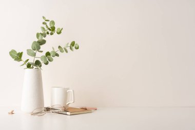İçinde beyaz bir vazo, okaliptüs, defter, gözlük ve kupa olan minimal bir masa kurulumu. Basitlik, organizasyon ve sakin bir çalışma ortamı için mükemmel bir kavram.