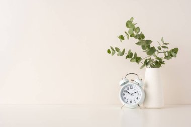 Yapraklı vazonun yanında klasik bir çalar saat içeren zarif minimalist kompozisyon.