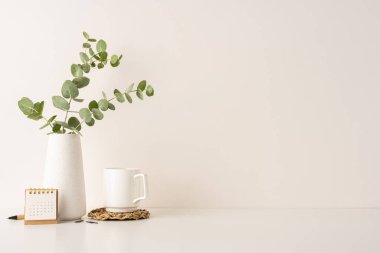 Beyaz bir vazoda okaliptüs, bir kahve fincanı ve küçük tahta bir takvimin yer aldığı modern ve temiz bir çalışma alanı. Minimalizmi, üretkenliği ve sakin bir çevreyi desteklemek için ideal.