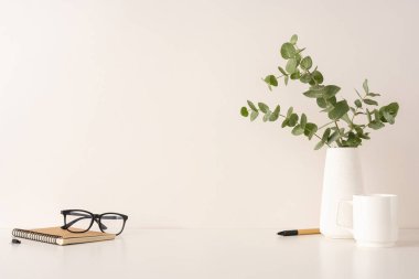 Beyaz bir yüzeyde dizüstü bilgisayar, gözlük, okaliptüs ve beyaz seramik eşyalar içeren sakin bir çalışma alanı düzenini gösteren bir kompozisyon, minimalist ve düzenli bir estetik sunar.