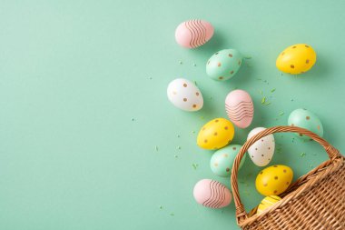 Yeşil bir yüzeyde sepetin yanına yerleştirilmiş renkli Paskalya yumurtaları, Paskalya kutlamalarının neşeli ve neşeli havasını somutlaştırıyor.