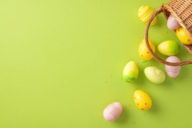 Renkli Paskalya yumurtaları, neşeli ve neşeli bir atmosfer yaratan bahar ve bayram kutlamalarını sembolize eden canlı yeşil bir arka planda hasır sepetin etrafına saçılmıştır.