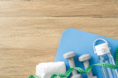 Spor salonu paspası, dambıl, havlu, su şişesi ve ahşap bir yüzey üzerinde ölçüm bandı gibi spor malzemeleri içeren düz bir düzenleme, sağlıklı bir yaşam tarzı ve ev çalışma rutinini vurguluyor.