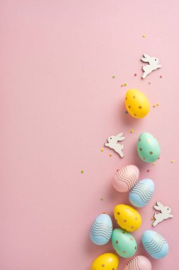 Parlak dikey Paskalya yumurtaları pembe bir yüzeye serilmiş. Dekoratif pozisyonlarda küçük ahşap tavşancıklar Paskalya mevsimi eğlencesini ve şenlikli süslerin yaratıcılığını kutluyorlar.