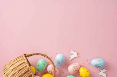 Renkli boyalı Paskalya yumurtaları ve pembe arka planda dekoratif olarak düzenlenmiş hasır bir sepet.