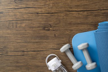 Mavi egzersiz paspası, dambıllar ve şişe tahta yüzeye yerleştirilmiş, fitness hedeflerini simgeliyor.