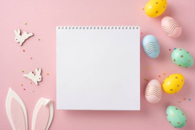 Paskalya yumurtaları, tavşanlar ve pembe arka plana karşı şenlikli dekorasyonlarla çevrili boş bir not defterinin üst görüntüsü, tatil temalı mesajlar veya yaratıcı notlar için yer sunuyor