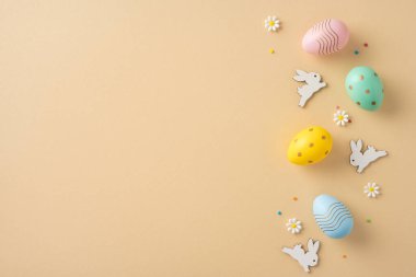 Canlı renkli Paskalya yumurtaları ve karışık tavşan süslemeleri bej bir yüzeye yayılmış, etrafı papatya ve konfetiyle çevrili.