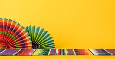 Çok renkli Meksikalı hayranlar ve seraptan oluşan dekoratif bir dekor, canlı sarı bir arka plan üzerine kurulmuş, festival ve kültürel bir ortam sağlıyor.