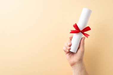 Eğitimi veya başarıyı sembolize eden kırmızı kurdeleyle bağlanmış bir diplomayı tutan bir elin yakınlaşması. Sade bej arka plan, diplomanın önemini ve zarafetini vurgular.