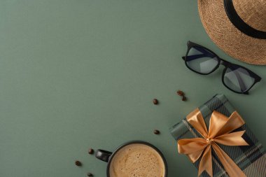 Bir kahve fincanı, altın kurdeleli yeşil bir hediye, şapka ve güneş gözlüğü içeren şık bir kompozisyon.