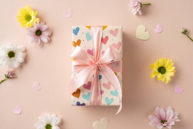 Dekoratif kalp temalı kağıda sarılı hediye kutusu, pembe bir kurdeleyle bağlanmış, çiçeklerle çevrili.
