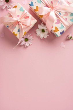 Beyaz papatyalar ve kalp aksanlarıyla çevrili pembe bir yüzeye yerleştirilmiş pembe kurdelelerle güzelce sarılmış dikey hediye kutuları.