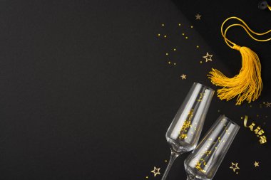 Mezuniyet temalı görüntüde iki şampanya kadehi, bir altın mezuniyet püskülleri ve şenlikli bir kutlama için dağınık konfeti yıldızları sergileniyor.