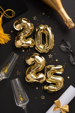 Mezuniyet şapkası, şampanya şişesi, altın aksanlar ve koyu dikey arka planda konfeti de dahil olmak üzere kutlama eşyalarıyla çevrili 2025 sayısını oluşturan altın balonların görüntüsü