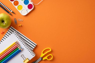 Okul malzemelerinin notlar, kalemler, makas, elma ve boya gibi yaratıcı düzenlemeleri canlı turuncu arka plan, eğitimi ve okula dönüş temalarını sembolize ediyor