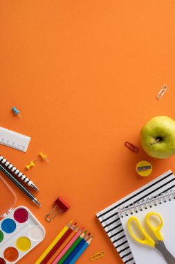 Kalemler, makas ve defterler de dahil olmak üzere renkli yatay okul malzemeleri, eğlenceli ve düzenli bir şekilde eğitime hazırlanmayı sembolize eden turuncu bir yüzey üzerinde sunulmaktadır.