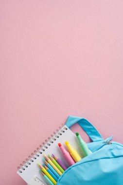 Renkli kalemler, işaretler ve pastel pembe bir yüzey üzerinde bir defter içeren bir sırt çantasının en üst dikey asgari görüntüsü