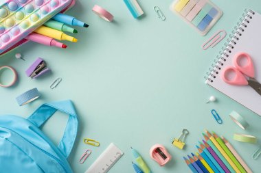 Pastel yeşil bir yüzey üzerinde çeşitli temel okul öğeleri düzenlenmiştir, renkli kırtasiye ve sırt çantası düzenlemeleri vurgulanır