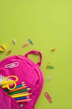 Yeşil bir yüzeye karşı pembe bir sırt çantasıyla düzenlenmiş dikey renkli okul malzemeleri.