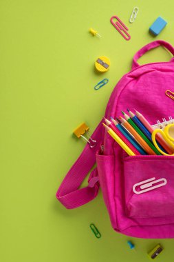 Okul malzemeleriyle dolu pembe bir sırt çantasının dikey düzenlenmesi. Kalemler ve makaslar da dahil. Canlı yeşil arka planda ek maddeler ile çevrili.