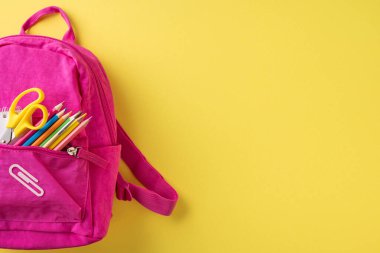 Parlak pembe okul sırt çantası. Katı sarı bir zemin üzerinde renkli malzemeler sergileniyor.