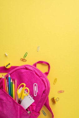 Okul malzemeleriyle dolu pembe dikey sırt çantası. Parlak sarı bir arka planda, yaratıcı bir şekilde düzenlenmiş.