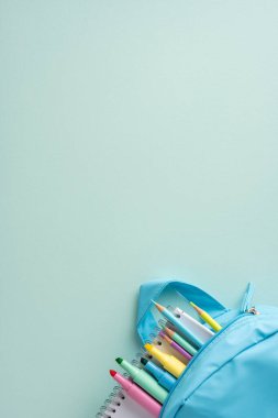 Minimalist bir pastel arka plana karşı kalemler ve defterler de dahil olmak üzere çeşitli renkli kağıtlarla dolu mavi düşey asgari bir sırt çantası.