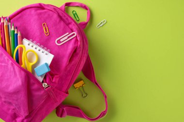 Canlı yeşil bir yüzeye yerleştirilmiş çeşitli okul malzemeleriyle dolu pembe bir sırt çantası.
