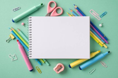 Kalemler, kalemler ve makaslarla çevrili boş spiral defter, pastel yeşil arka planda.